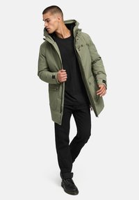 Olive zelena parka s kapuco, sprednjim zadrgo in več žepi; kombinirana z črnima majico in temnimi hlačami; gladka tekstura tkanine.