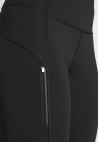 Rugalmas anyagból készült fekete leggings, tónusos varrással, oldalsó cipzáras zsebbel és szoros, testhezálló kialakítással.
