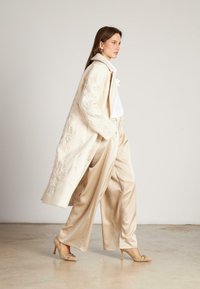 Largo abrigo de crema con bordado floral, combinado con pantalones de pierna ancha beige brillantes y zapatos de tacón de color claro. Fondo neutro.