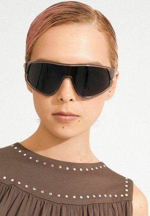 Črna prevelika sončna očala z geometričnim dizajnom, z debelim prozornim rjavim okvirjem in temnimi lečami, nošena z rjavim zgornjim delom z zakovicami.