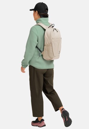 Persona con cappello nero, giacca in pile verde, pantaloni marroni e scarpe da trekking rosa e nere che cammina, con zaino beige su sfondo chiaro.