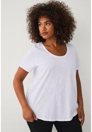 Kvinde med krøllet hår, iført øreringe i hoops, en hvid T-shirt med korte ærmer og sorte bukser, der kigger mod venstre mod en ensfarvet baggrund.