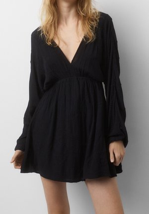 Femme portant une robe mini noire ample à manches longues, avec un décolleté en V profond et une taille élastique, debout devant un fond clair uni.