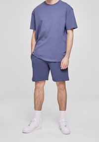Uomo che indossa una camicia blu a maniche corte e pantaloni corti abbinati, con calzini bianchi e sneakers bianche, in piedi davanti a uno sfondo semplice.