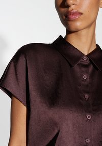 Chemise bordeaux foncé en tissu texturé, à manches courtes, avec un col et des boutons assortis. La vue en gros plan se concentre sur la partie supérieure.