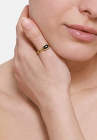 Gouden ring met een rechthoekige zwarte steen, met een gedraaide band en een gedetailleerde rand rond de steen. Hand rustend tegen een nek.