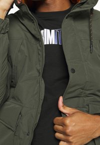 Giacca in nylon verde scuro con zip frontale e bottoni a scatto, dotata di cappuccio, due tasche laterali e polsini elasticizzati. Maglietta nera visibile sotto.