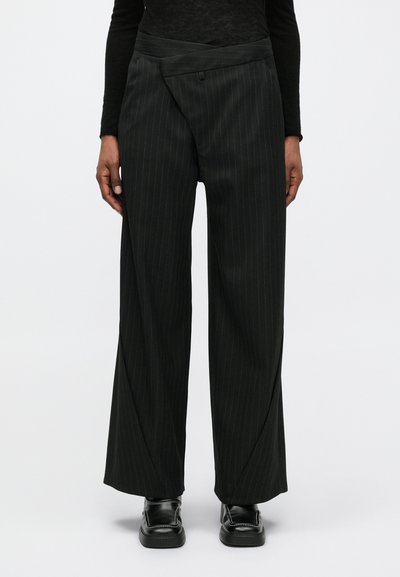 Pantalons larges à rayures noires avec une taille asymétrique, portés avec un top noir à manches longues et des mocassins noirs.