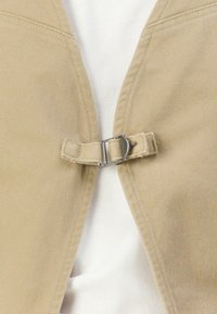 Beige tekstilrem med sølvfarget metallspenne som fester to beigematerialer over en hvit tekstil bakgrunn.