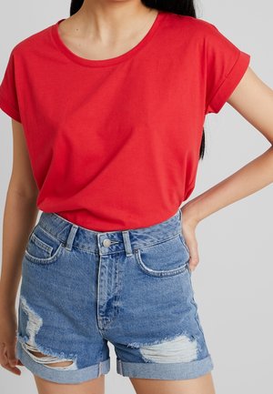 T-shirt in cotone rosso con maniche corte arrotolate, abbinato a pantaloni corti in denim blu a vita alta con bordi sfrangiati e dettagli usurati.