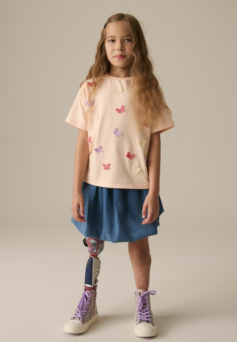 Helles rosa T-Shirt mit buntem Schmetterlings-Stickerei, kombiniert mit einem Jeansrock. Verfügt über ein buntes Prothesebein und glitzernde High-Top-Schuhe.