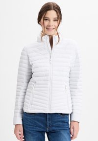 Weiße Daunenjacke mit horizontaler Steppstruktur, hohem Kragen und mit Reißverschlüssen versehenen Taschen, kombiniert mit blauen Jeans.