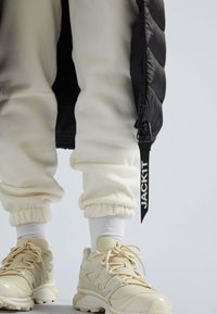 Beige atletiksko med strukturerede såler, cremefarvede afslappede sweatpants med samlede manchetter og en sort polstret jakke med lynlås.