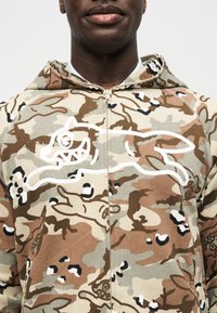 Isik, kes kannab kamuflaažiga lukuga dressipluusi, millel on valge kontuuriga koomiksikoer rinna kohal ja samas mustris kapuuts.