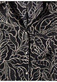 Sort bluse med et beige bladmønster, der har knapper, krave og tekstureret stof. Bemærkelsesværdige detaljer inkluderer intrikate linjearbejder i designet.