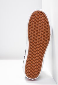 Sneaker mit weißem Obermaterial und einer strukturierten Gummisohle in Hellbraun, die ein diagonales Muster für besseren Halt aufweist.