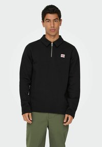 Sweatshirt noir à col zippé, manches longues, avec un petit patch logo rouge et blanc sur la poitrine, associé à un pantalon vert clair.