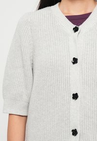 Cardigan en maille gris clair à manches courtes avec des boutons noirs en forme de fleur porté sur un top violet.
