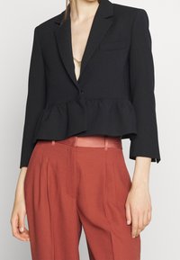 Le mannequin porte un blazer noir à basque sur un top beige, assorti à un pantalon taille haute couleur rouille, mettant en valeur une tenue élégante.