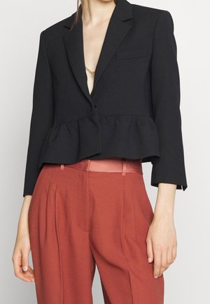 Il modello indossa un blazer peplum nero sopra un top beige, abbinato a pantaloni a vita alta color ruggine, mostrando un look elegante.