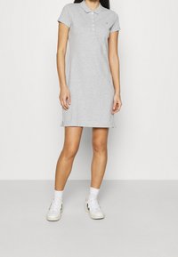 Grijze polojurk met korte mouwen en een knoopsluiting. Bevat zijsplitten en een klein logo detail. G gedragen met witte sneakers en sokken.