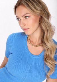 Pruhovaný modrý top s krátkým rukávem, který má kulatý výstřih a texturovaný vzor. Modelka nosí zlatý řetízek.