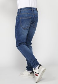 Jeans de mezclilla azul ajustados con diseño de cinco bolsillos, costuras en contraste y una etiqueta de cuero en la parte posterior de la cinturilla. Combinados con zapatillas blancas.