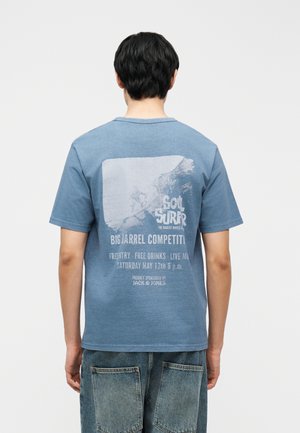 Man wearing blue t-shirt with faded surfing contest graphic and text on the back, paired with loose denim shorts.