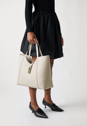 Borsa a tracolla in pelle beige con doppi manici, dotata di una chiusura dorata e dettagli di cucitura verticale. Indossata con una gonna nera e scarpe nere.