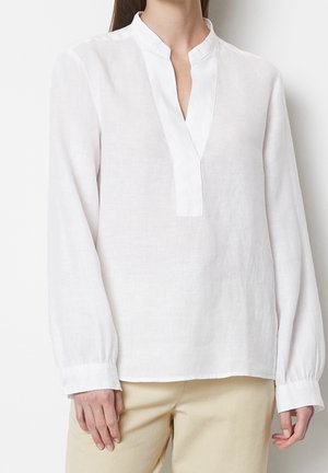 Femme portant un chemisier en lin blanc à manches longues avec un col mao et un pantalon beige, debout devant un fond uni.