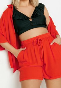 Czarny top w falbanki z dekoltem w kształcie łezki, zestawiony z luźnymi czerwonymi szortami z elastycznym pasem i bocznymi kieszeniami. Czerwony narzutka na ramionach.