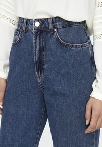 Högmidjade blå denimjeans med kontrasterande sömmar, fem fickor och knappstängning framtill. Tyget har en lätt textur och lyster.