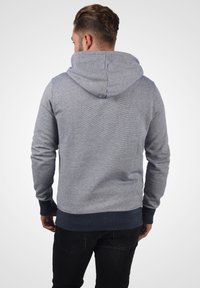 Grauer Kapuzenpullover mit einem strukturierten Design, ausgestattet mit einer Kordelzugkapuze, gerippten Bündchen und einem dunkelblauen Saum. Sichtbare Nähte entlang der Nähte.