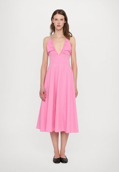 Robe rose à bretelles avec un décolleté en V profond, des détails froncés et une jupe évasée fluide. Stylisée avec des ballerines noires sur un fond neutre.