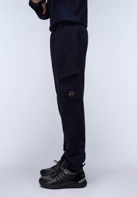 Navy sweatpants with a side pocket and a patch logo, made of soft fabric. Paired with black athletic shoes featuring gray accents.