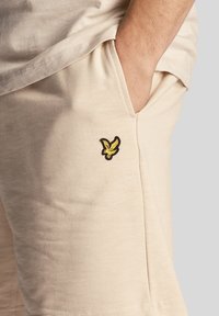 Beige Shorts aus einem weichen, strukturierten Stoff mit seitlichen Taschen und einem kleinen, schwarz-gelben gestickten Logo am linken Bein.