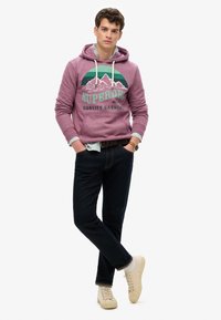 Fioletowa bluza z kapturem z górskim nadrukiem i napisem "Superdry", w połączeniu z ciemnymi dżinsami i beżowymi sneakersami. Widoczne sznurki i warstwowy wygląd.