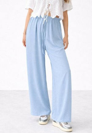 Femme portant un pantalon ample bleu clair avec taille à cordon, un haut blanc à lacets et des baskets compensées argent-blanc, debout sur un sol clair.