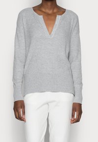 Gray ribbed knit sweater with a V-neck, dropped shoulders, and long sleeves, featuring a subtle label near the neckline. Paired with white pants.