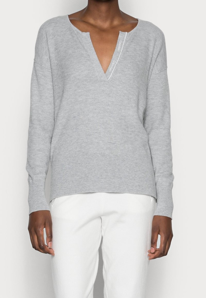 Gray ribbed knit sweater with a V-neck, dropped shoulders, and long sleeves, featuring a subtle label near the neckline. Paired with white pants.