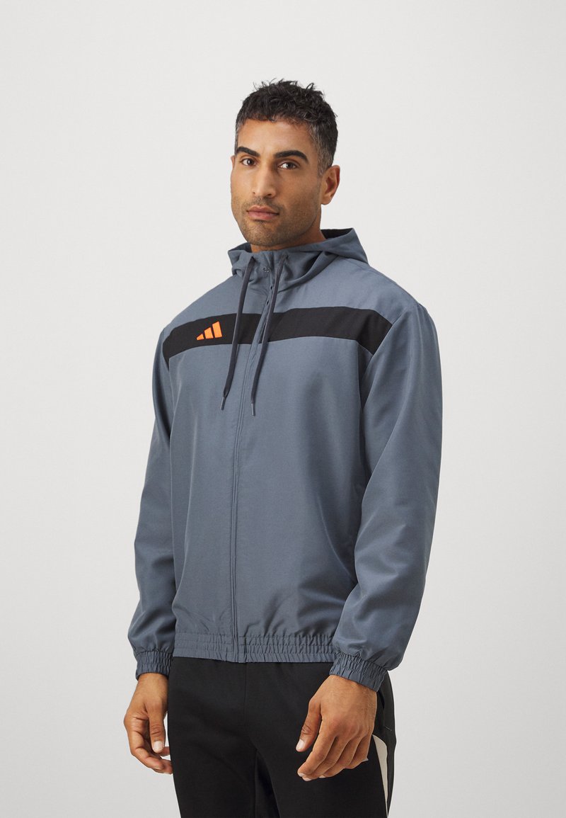 Grå vindjakke med en sort vandret stripe og orange detaljer, med hætte, lynlås foran og elastiske manchetter.