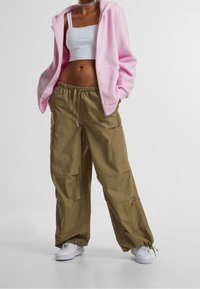 Sudadera con capucha de color rosa claro con cremallera frontal, llevada sobre una camiseta de tirantes blanca. Pantalones cargo verde oliva con bolsillos y puños elásticos. Zapatillas blancas.