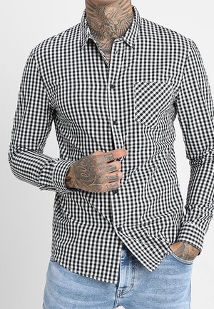 Chemise à carreaux noir et blanc avec boutons, portée par une personne tatouée, une main ajustant la chemise et l’autre dans la poche d’un jean bleu.