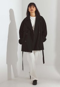 Cappotto trench nero doppiopetto con ampi risvolti, tasche frontali e cintura; abbinato a pantaloni bianchi e scarpe nere. Texture liscia, vestibilità sartoriale.