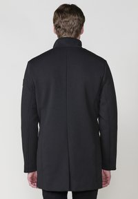 Abrigo negro ajustado con un cuello alto, textura lisa similar a la lana y silueta estructurada. Presenta líneas limpias y detalles de costura mínimos.