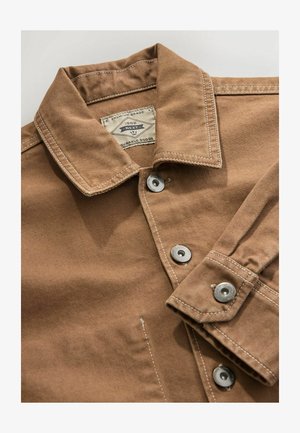 Chemise en denim brun avec un col, des boutons pressions argentés et des coutures blanches contrastantes. L'étiquette indique "Next Durable Goods."