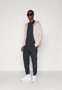 Letvægts beige jakke med hætte, sort T-shirt og sorte træningsbukser. Har hvide detaljer og Nike-logoer. Hvide atletiske sko fuldender looket.