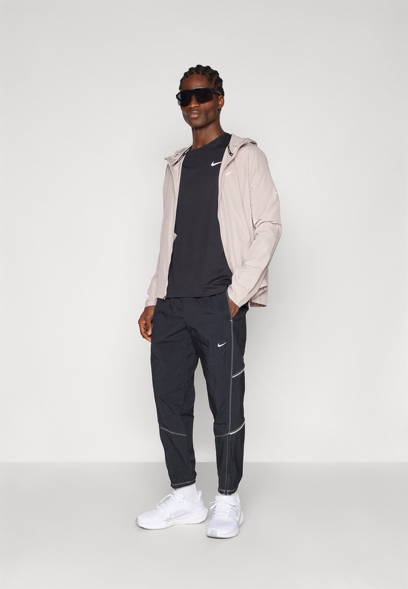 Leichte, beige Jacke mit Kapuze, schwarzes T-Shirt und schwarze Sporthosen. Mit weißen Akzenten und Nike-Logos. Weiße Sportschuhe runden das Outfit ab.