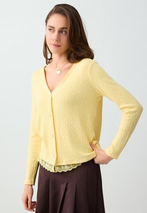 Jonge vrouw die een lichtgele gestructureerde cardigan draagt over een top met kanten rand en een donkerbruine rok, kijkt opzij met hand in de zij.