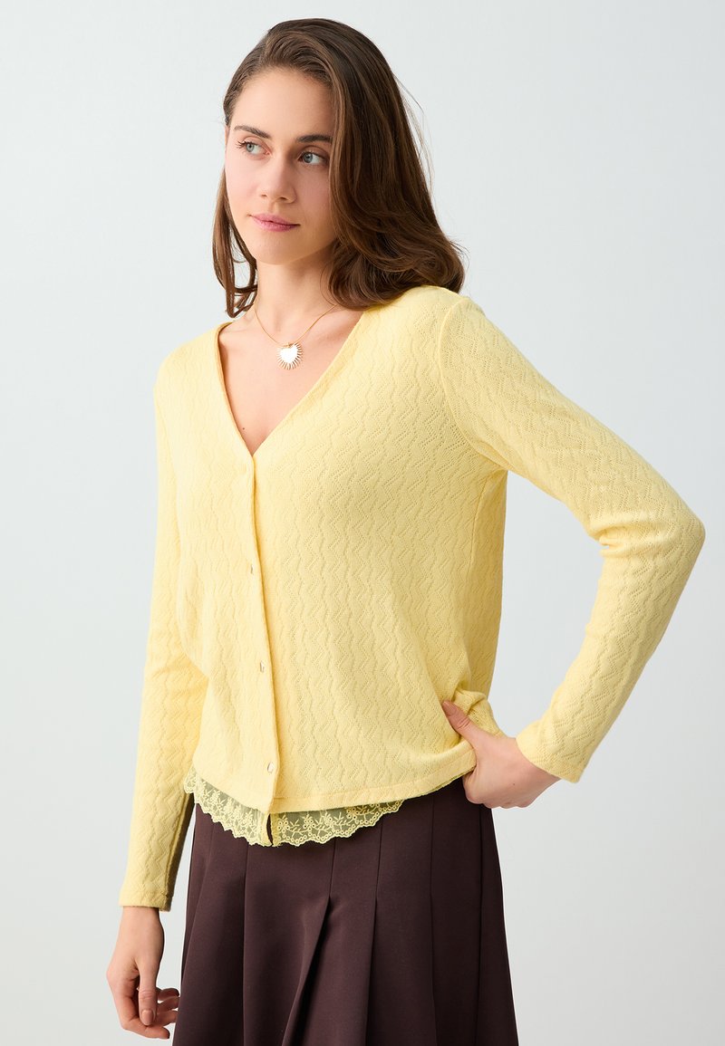 Jeune femme portant un cardigan texturé jaune pâle sur un haut bordé de dentelle et une jupe marron foncé, regardant sur le côté avec la main sur la hanche.